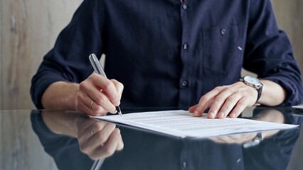 Businessman in dark blue t-shirt is signing contract at desk. Close-up of a male executive signing...