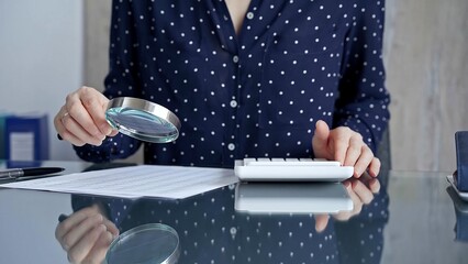 Auditor woman analyzing financial documents with magnifying glass and calculator at her office desk in formal dark blue blouse with polka dots. Business people concept