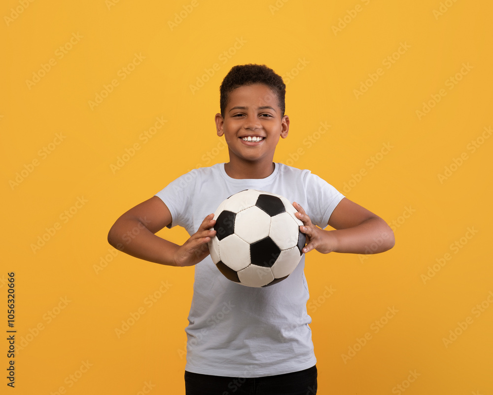 Wall mural cheerful black school boy holding soccer ball and smiling at camera, playing football, yellow studio