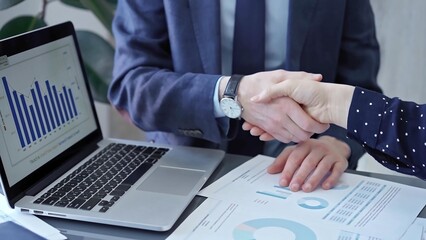 Two business people professionals shaking hands in office setting, sealing a deal with laptop and financial graphs visible. Business handshake over laptop and reports