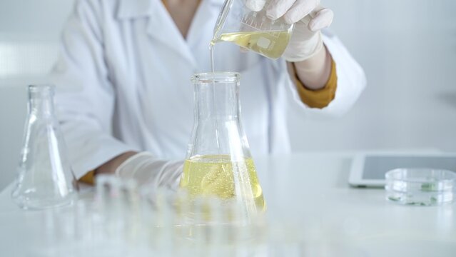 A scientist, wearing a lab coat and white protective gloves, is pouring a yellow oily liquid from one beaker to another in laboratory, close up. Medicine and science concept