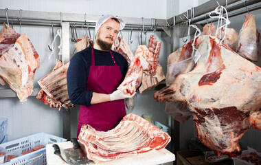 Skilled positive young bearded butchery worker standing at cutting table in meat processing room,...
