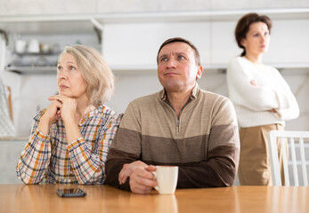 Livid middle-aged man sitting at the kitchen-table next to old female family member looking upset