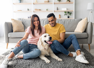 Full length portrait of cheery multiracial couple with their pet dog sitting on floor at home. Arab...
