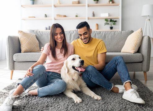 Fototapeta Full length portrait of cheery multiracial couple with their pet dog sitting on floor at home. Arab man and his Caucasian wife spending time with golden retriever in living room