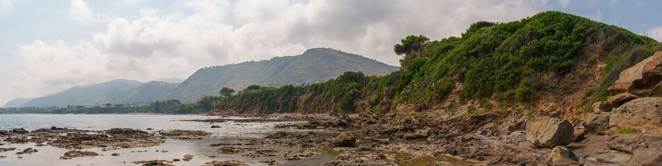 A wide-angle panoramic view of a rugged coastal landscape featuring rocky shores, lush green cliffs, and distant hills under a partly cloudy sky. Ideal for nature and travel photography