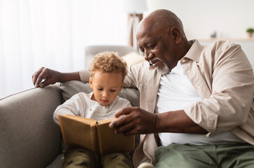 African American Grandpa And Grandson Reading A Book Spending Time Together Sitting On Couch At Home On Weekend. Senior Grandfather Teaching Grandchild To Read Indoors. Knowledge, Family Leisure