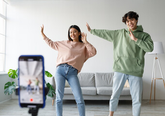 Young Asian couple recording video content, dancing on smartphone camera at home. Positive millennial boyfriend and girlfriend filming for social media, using mobile device. Vlogging concept