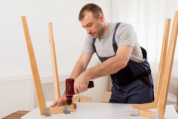 Male Carpenter Using Electric Drill Assembling Wooden Table After Transportation To New Apartment. Man In Blue Coverall Uniform Fixing Desk With Carpentry Tools Working In Studio.