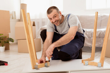 Happy Carpenter Male Assembling Table After Transportation Smiling To Camera Working Indoors, Wearing Coverall Uniform. Furniture Assembly And Carpentry Service Concept