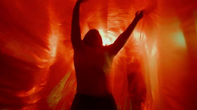 Expressive shirtless dancer performing graceful, dynamic body movements between translucent plastic curtains illuminated by dramatic colorful lighting. Low angle view