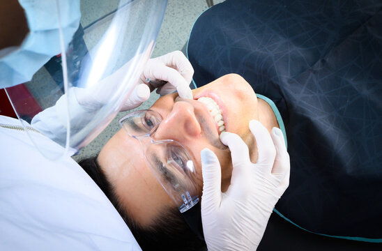 A patient lies back in a dental chair while a dentist inspects their teeth, using specialized tools and wearing protective gear in a clean, well-lit office.