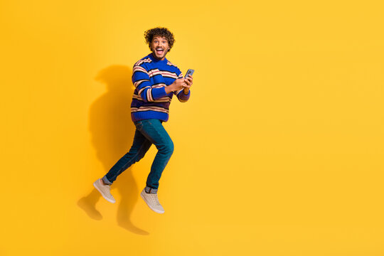 Full length photo of handsome young guy running hold gadget dressed stylish blue striped garment isolated on yellow color background
