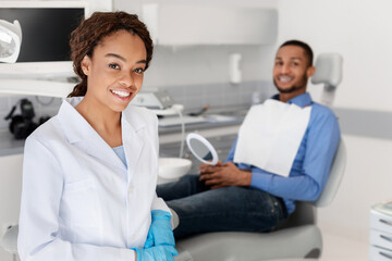 Fototapeta premium Smiling african woman dentist and young man patient looking at camera, dental cabinet interior