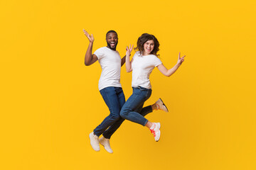 Playful multiracial spouses having fun, jumping in the air over yellow studio background, woman...