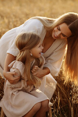 Little blonde girl in beige dress hugs her mother in the rays of warm summer sunset. Happy smiling...
