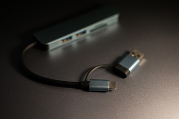 USB-C hub in gray, featuring multiple USB ports and card reader slots, with attached USB-C and USB-A connectors, resting on a dark metallic surface. Compact connectivity solution.