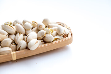 A light-colored basket filled with pistachio nuts, some scattered on a white surface. The pistachios, in their shells with some slightly open, reveal the green nut inside, showcasing a nutritious snac
