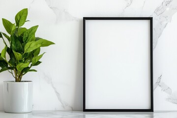 A stylish black-framed blank picture frame beside a lush green plant on a marble countertop.