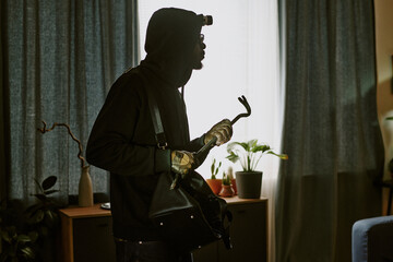 Side view of African American thief in total black wearing dirty leather gloves and holding metal...
