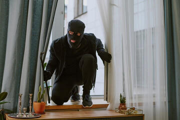 Man in total black with balaclava and crowbar sitting on windowsill after break-in