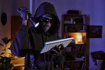 African American in total black clothes with hood looking at wireless keyboard in dark room