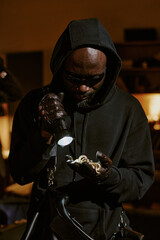 Vertical shot of African American thief using flashlight to illuminate golden jewelry in hands