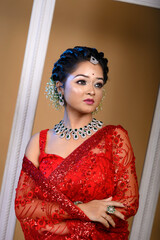 Portrait of beautiful young Indian woman wearing a traditional red saree adorned with delicate jewellery posing fashionably in studio lighting