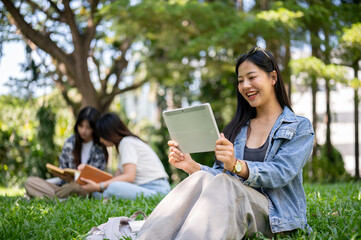 A beautiful Asian female college student sits on grass in a park, using her digital tablet.