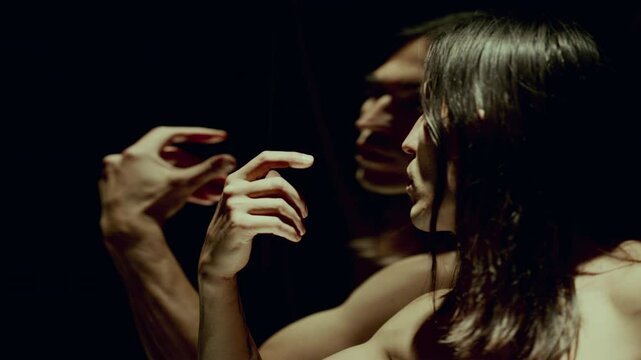 Shirtless male dancer performing enigmatic moves in dark studio with dramatic lighting, looking at his warped reflection in distorting mirror