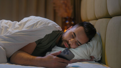 Caucasian man lying in bed bedroom user mobile phone smartphone cellphone gadget technology male guy connected scrolling website browsing network cyber ecommerce chatting internet distance communicate