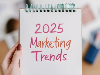 A hand holding a notebook with the words "2025 Marketing Trends" written in colorful text, symbolizing future marketing strategies and innovations.