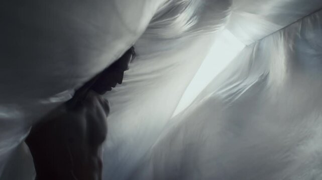 Silhouetted dancer with long hair and shirtless torso moving gracefully between flowing translucent plastic curtains, creating hypnotic contemporary performance in backlit studio