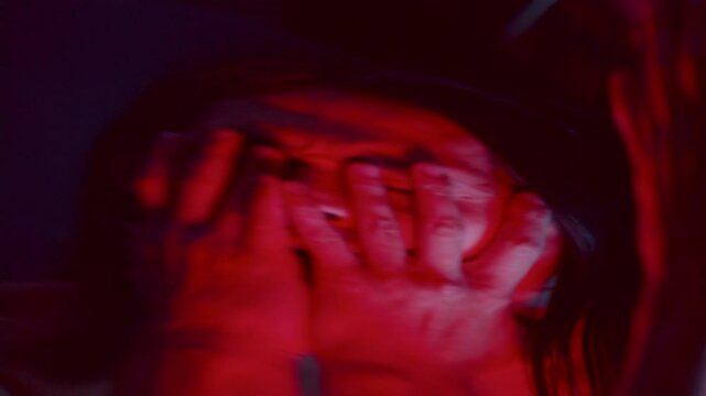 Warped reflection in distorting mirror of frightened or desperate man covering his face with hands in fear in dark studio with red lighting