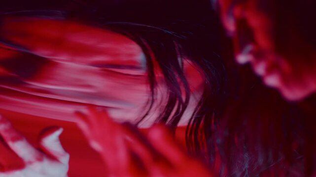 Bewildered man with long hair examining his palms in front of a distorting mirror that surreally warps his reflection in dark studio with red illumination