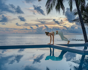 Young woman in a white clothes doing yoga on the beach near swimming pool early morning in the summer. Sunrise sky in the background .	

