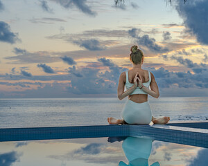 Young woman in a white clothes doing yoga on the beach near swimming pool early morning in the summer. Sunrise sky in the background .	
