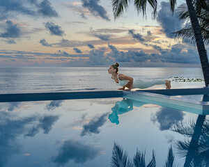 Young woman in a white clothes doing yoga on the beach near swimming pool early morning in the summer. Sunrise sky in the background .	
