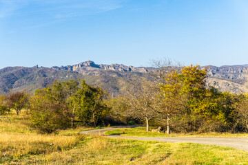 Earth road, grass and trees with autumn leaves. Mountain with ruins of Birtvisi fortress, bright sky in the background