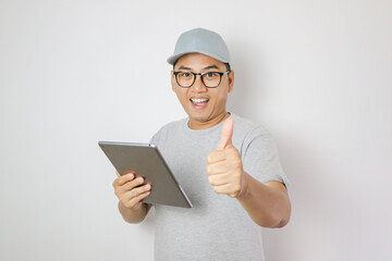 Asian delivery man or courier smiles and gives a thumbs up gesture to the camera while peeking out from behind a white wall. Concept of delivery services, customer satisfaction, business reliability