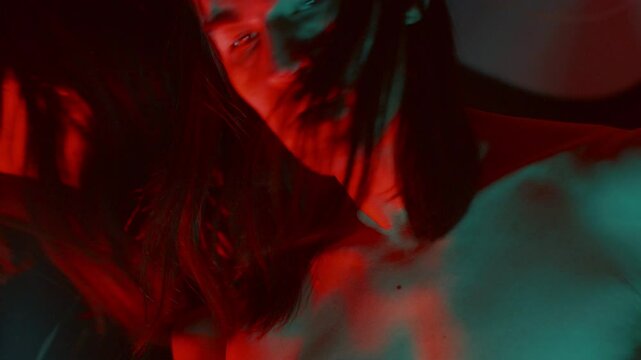 Long-haired man with shirtless torso looking around confusedly in front of distorting mirror in dark studio with red and blue neon illumination