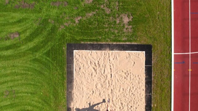 Aerial view of an athlete landing in a sandpit after a long jump, her shadow stretched out across the sand under bright sunligh