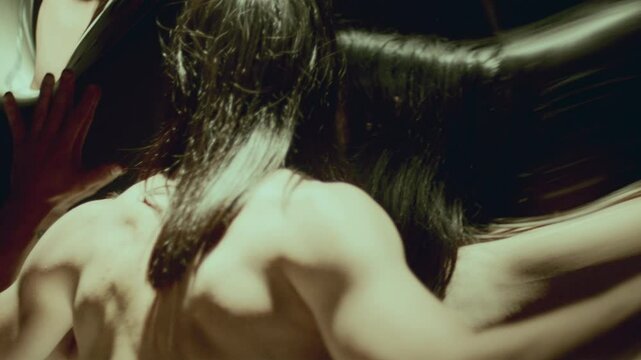 Fascinated shirtless man with long hair staring intently into distorting mirror, holding his hands on reflective surface in dark studio