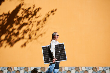 Woman holding solar panel for charging, stands against vibrant orange wall. Smiling girl wears...