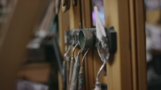 No people closeup of row of palette paint scrapers and spatulas, hanging on magnetic strip below wooden easel in tiny artist studio in ambient day light