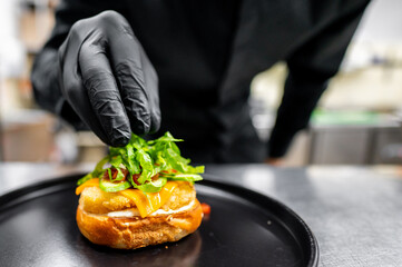 A close-up of a hand in a black glove placing a gourmet burger with cheese and greens on a black plate, emphasizing freshness and quality. Perfect for culinary blogs, and restaurant promotions.
