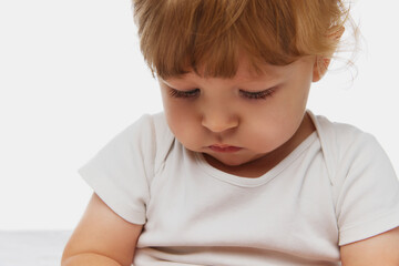 Close-up of baby looking down with thoughtful expression, dressed in white onesie against white studio background. Concept of parenthood, natural beauty, love and care. Ad.