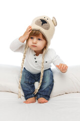 Baby dressed in cozy bear hat and jeans, touching hat while seated on bed against white studio background. Kid's fashion. Concept of parenthood, natural beauty, love and care. Ad.