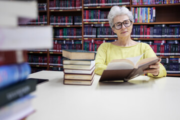 Senior woman reading a book in a library, surrounded by stacks of books, wearing glasses, focused...