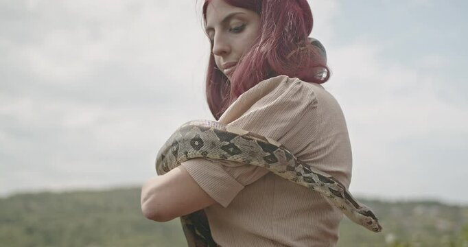 A young woman with red hair shows affection to her pet snake on a sunny day.